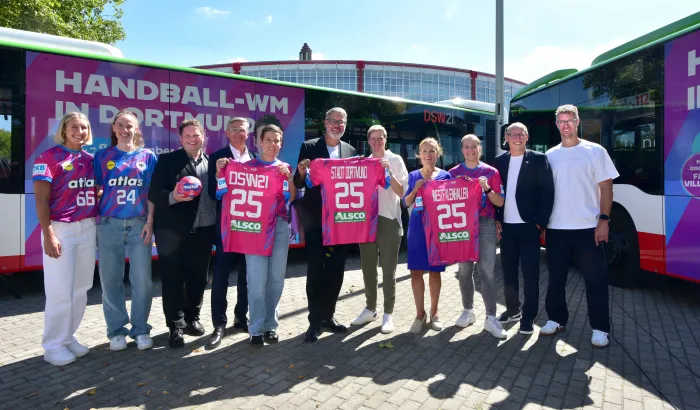 Spielerinnen und Verantwortliche von DHB, DSW21, Westfalenhallen und der Stadt Dortmund posieren mit Trikots und dem WM-Ball vor einem Bus im WM-Design
