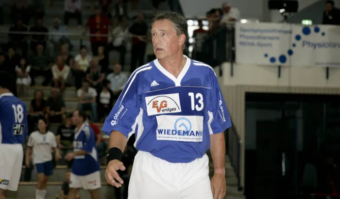 Ein älterer Mann in einem blauen Trikot der in einer Handballhalle steht.