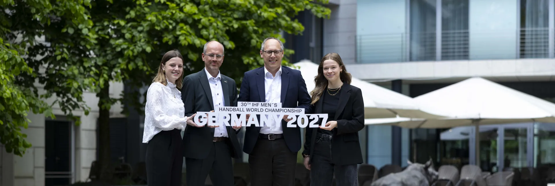 Zwei Männer und zwei Frauen halten vor einem Radisson Hotel den Schriftzug der Handball-Weltmeisterschaft 2027 in die Höhe.