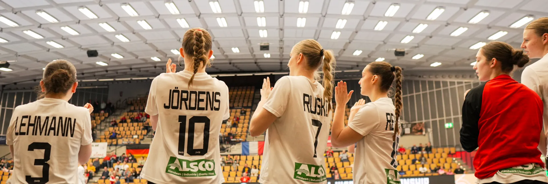 Eine Truppe von Handballerinnen in weißen Trikots die bei einem Tor aufstehen und jubeln