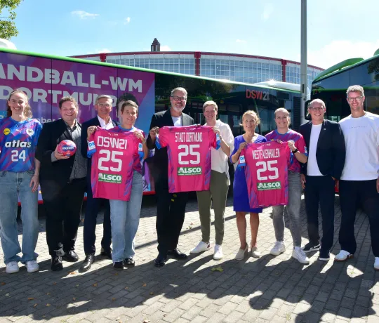 Spielerinnen und Verantwortliche von DHB, DSW21, Westfalenhallen und der Stadt Dortmund posieren mit Trikots und dem WM-Ball vor einem Bus im WM-Design