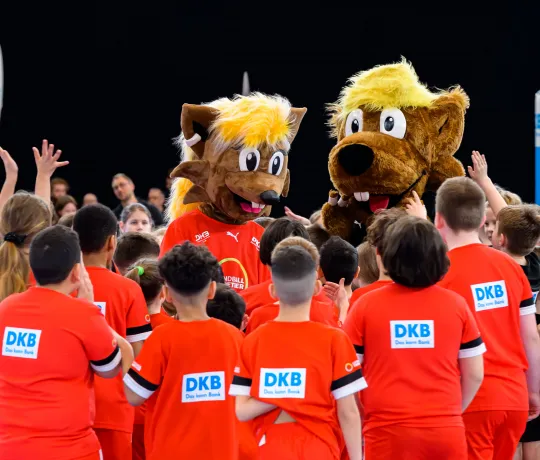 Viele Kinder stehen um zwei Maskottchen herum und jubeln ihnen zu. 