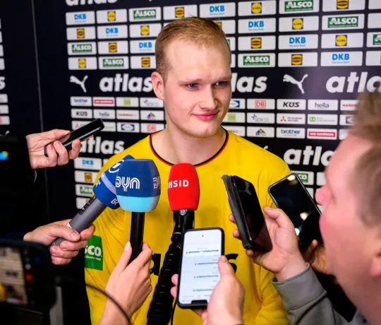 Ein Handballspieler im Interview, zu ihm werden viele Aufnahmegeräte gehalten, in die er hinein spricht. 
