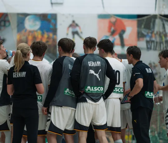 Eine Truppe von Handballern in weißen Trikots die bei einem Timeout im Kreis zusammenstehen 