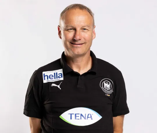 Ein Mann mit verschränkten Armen hinter dem Rücken der in die Kamera lächelt.