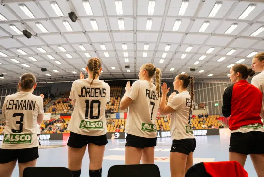 Eine Truppe von Handballerinnen in weißen Trikots die bei einem Tor aufstehen und jubeln